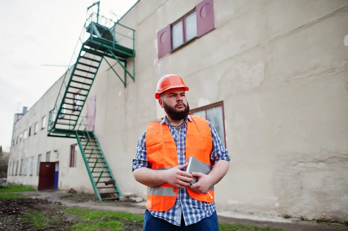 A tough, bearded man in a suit, a construction worker wearing a safety orange helmet, stands near large industrial stairs.