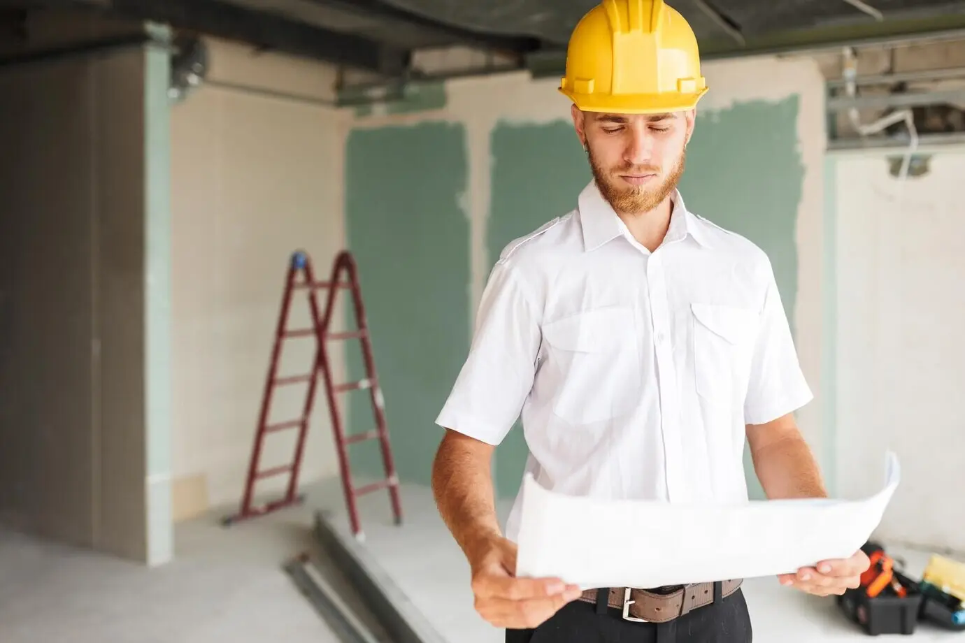 A young architect wearing a white shirt and a yellow hardhat is thoughtfully looking at the plan of a new apartment, with a ladder in the background.