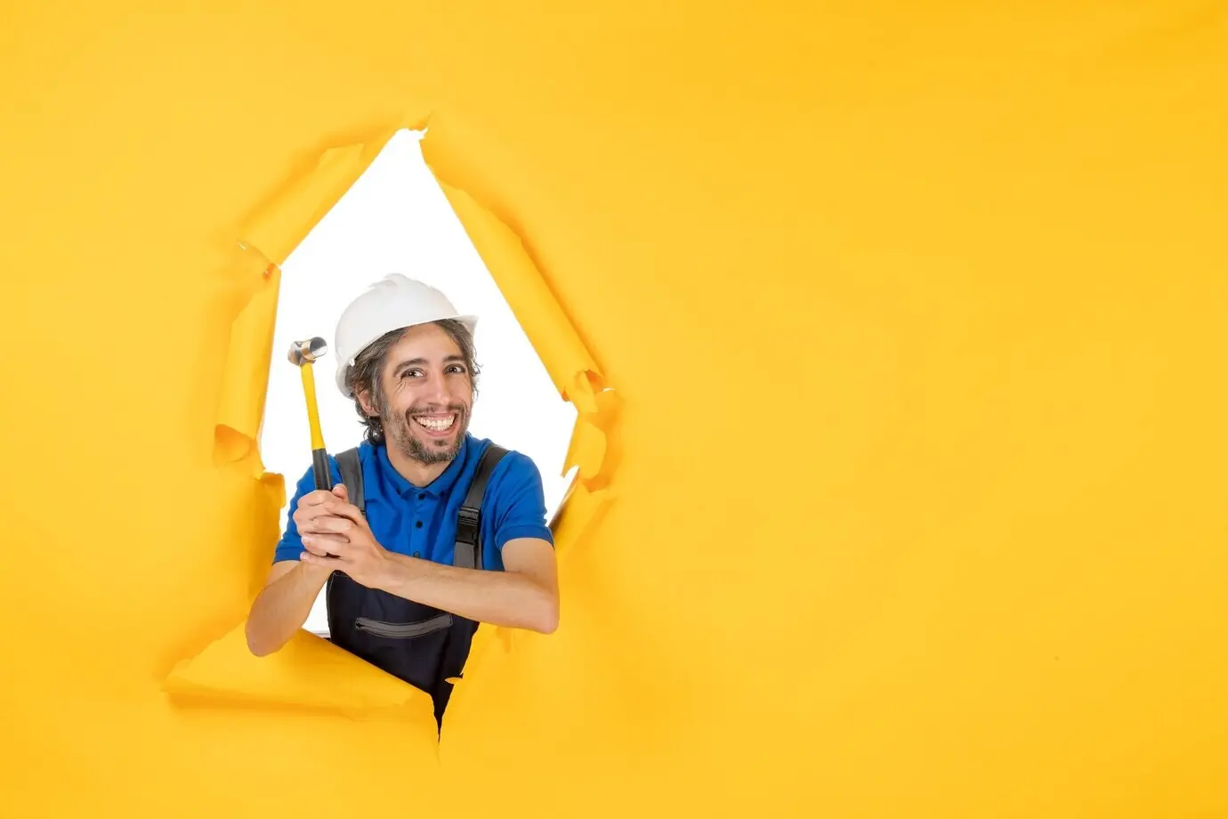 Frontal view of a male builder in uniform holding a hammer against a yellow wall; worker man, building job, constructor, architecture, color.