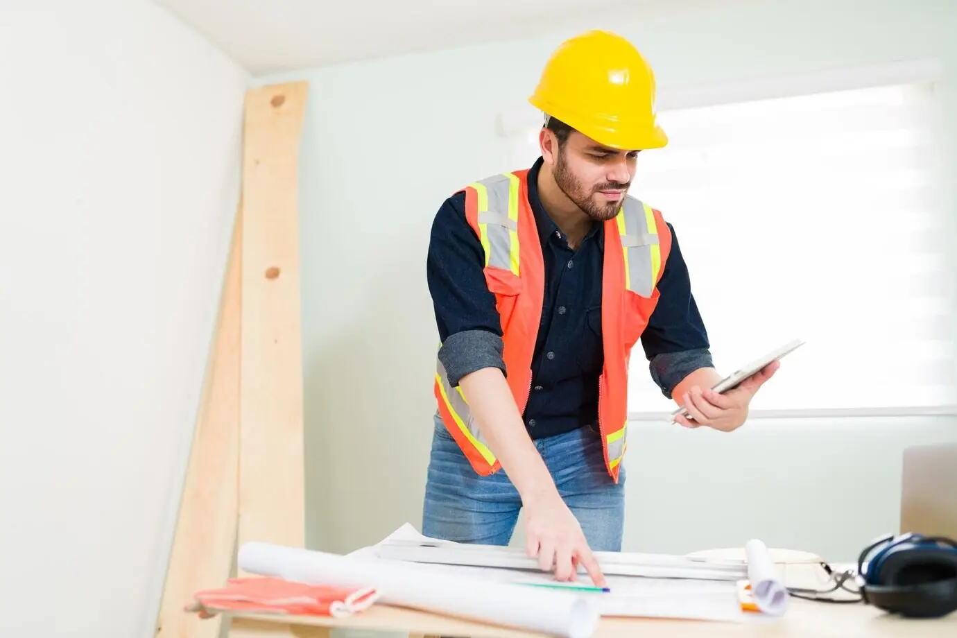 A handsome man and a contractor wearing a security helmet review the construction blueprints while looking at a tablet.
